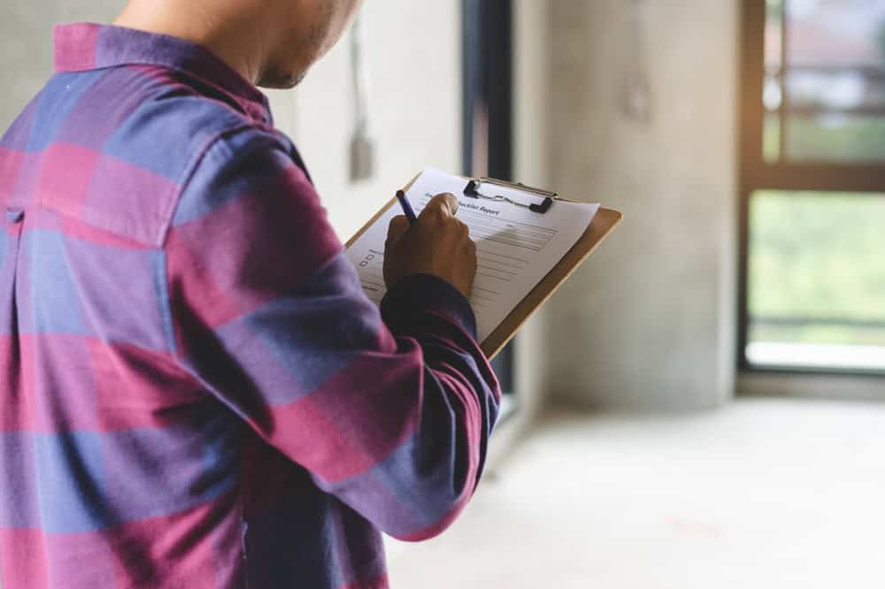 A person in a plaid shirt holds a clipboard and writes on a form, perhaps conducting lead paint testing. The background reveals a window and an unfinished room with concrete walls, typical scenes for home inspectors across Long Island.