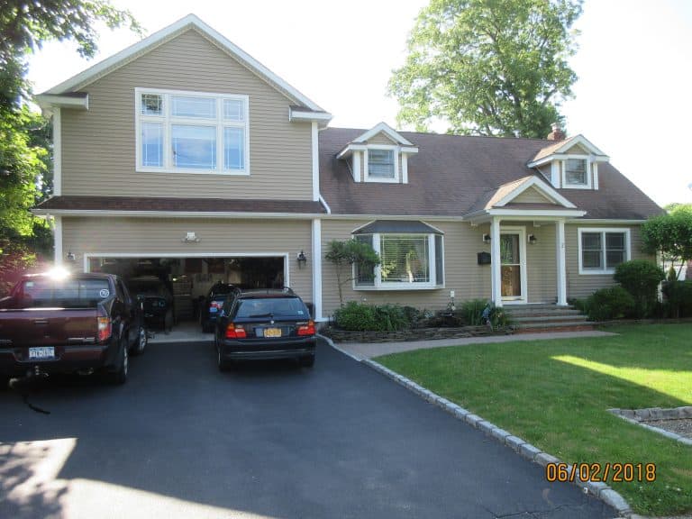 A suburban house with beige siding and a gable roof stands quietly. Two cars rest in the driveway, while the garage door opens to reveal another car inside. A large tree and landscaping enhance the view, dated '06/02/2018,' ideal for Home Inspections Long Island looking to capture every detail.