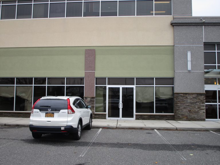 A white SUV is parked in front of a modern commercial building with large windows and a varied facade of beige, green, and gray tones. The empty parking lot hints at the focus on detail typical during Commercial Inspections on Long Island.