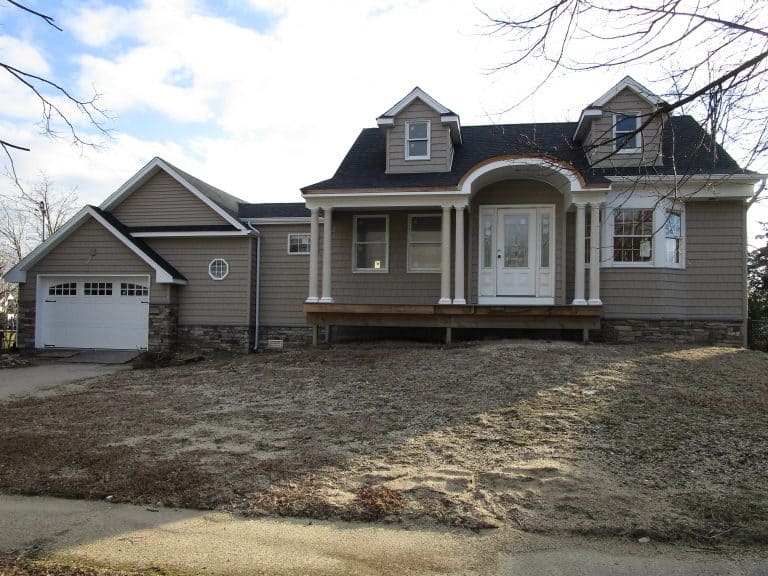 A two-story house with a front porch and bay windows, gray siding, black roof, and a garage on the left sits quietly. The yard is bare with sparse grass and leafless trees under a partly cloudy sky. Perfectly suited for a detailed evaluation by a home inspector on Long Island.