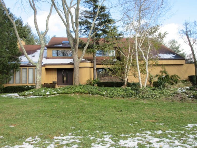 A tan and brown two-story house with a sloped roof, ideal for Home Inspections Long Island, is surrounded by trees. Snow dusts the ground with patches of grass in the front yard. The sky is partly cloudy.