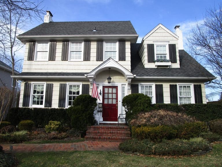 A two-story white house with a dark gray roof and shutters, and a red front door graces Long Island's welcoming charm. A small porch invites you in from the landscaped yard, where meticulous home inspections ensure safety and peace of mind. A brick pathway gleams under the clear, sunny sky.