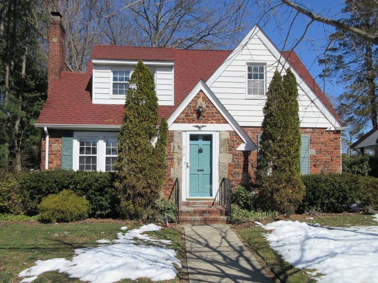 A charming two-story brick house with a red roof and a teal front door, perfect for a thorough home inspection on Long Island. Flanked by tall evergreen shrubs, it stands amidst a lawn with patches of snow, against a backdrop of leafless trees and a clear blue sky.