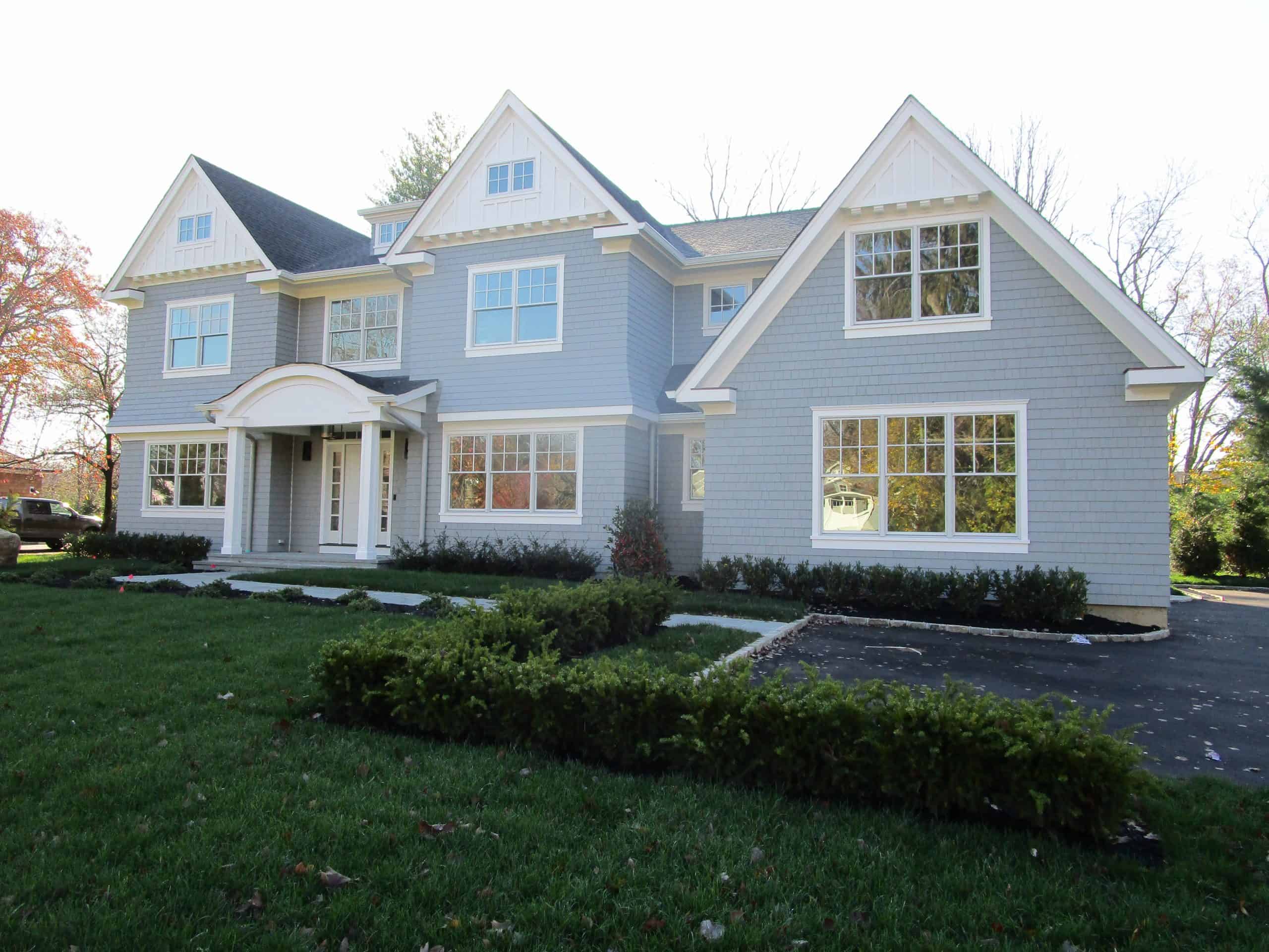 A two-story house with a gray exterior and white trim showcases multiple gabled roofs. Large windows and a covered entrance add charm, while a manicured lawn and hedge line the driveway. Ideal for an expert review, it's perfect for home inspections on Long Island. Trees grace the background elegantly.