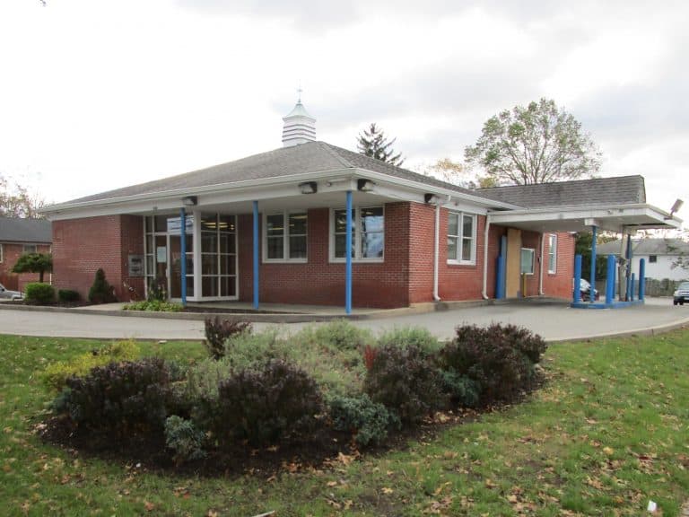 A small, red brick building with a drive-through area and blue columns stands resilient under a gray, cloudy sky. In the foreground, a garden with shrubs and grass adds charm. Perfect for those considering commercial inspections on Long Island to evaluate such inviting spaces.