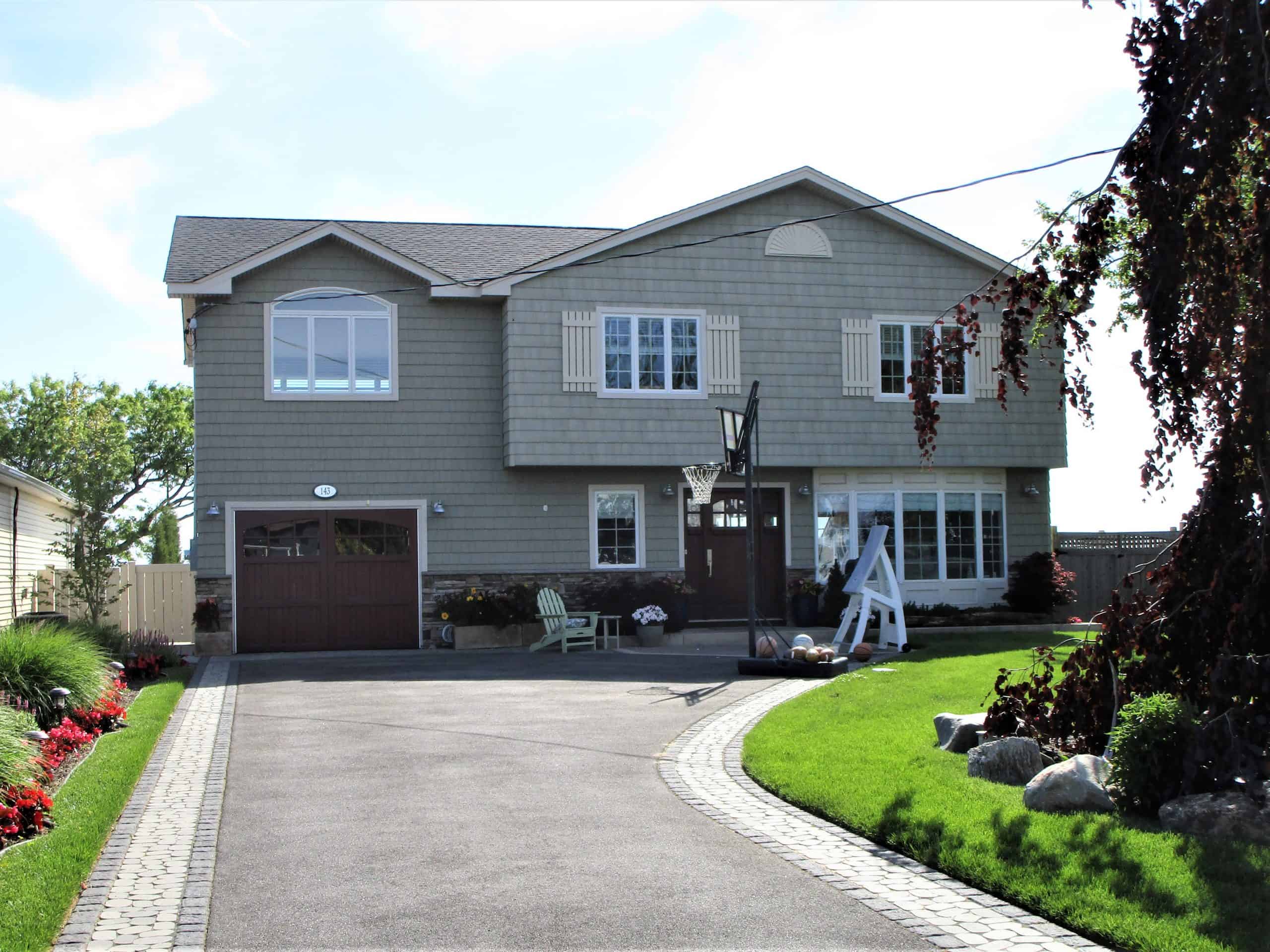 A two-story house with gray siding, large windows, and a garage awaits. A paved driveway leads to the entrance, surrounded by a lush lawn and flowers. A white decorative anchor sits near the front, adding charm. Perfect for those scheduling home inspections on Long Island to ensure peace of mind.