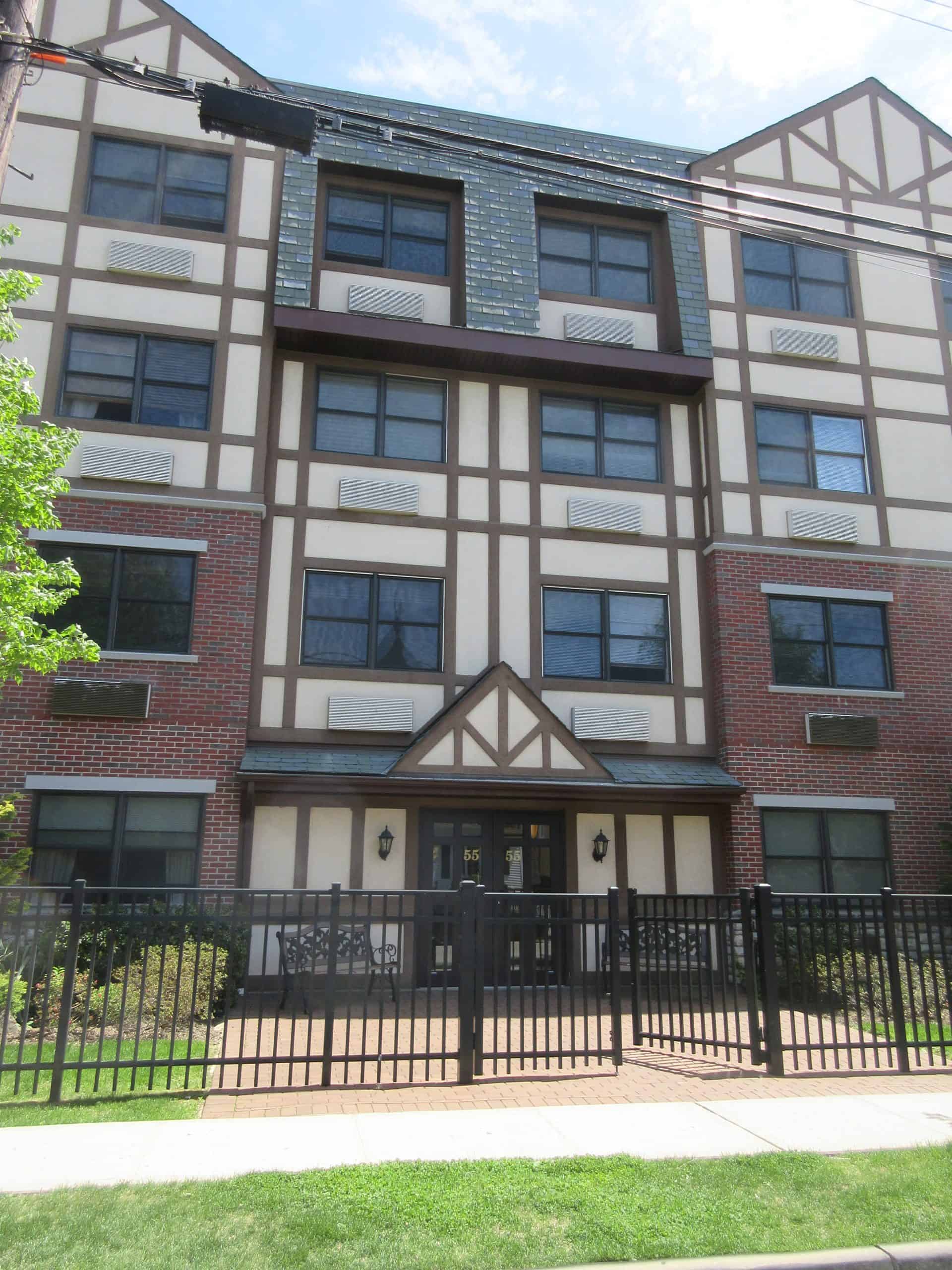 A four-story building with a Tudor-style facade, featuring timber framing and brickwork, awaits your visit. With the added assurance of Home Inspections Long Island, the property boasts large windows on each floor. A gated entrance is bordered by a green lawn for picturesque charm.