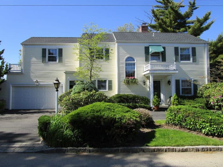 A two-story white house with green shutters and a small front porch offers charm, while the manicured front yard adds appeal. Ideal for those considering home inspections in Long Island, the driveway leads to a left-side garage, and a lamppost stands tall against the clear blue sky.