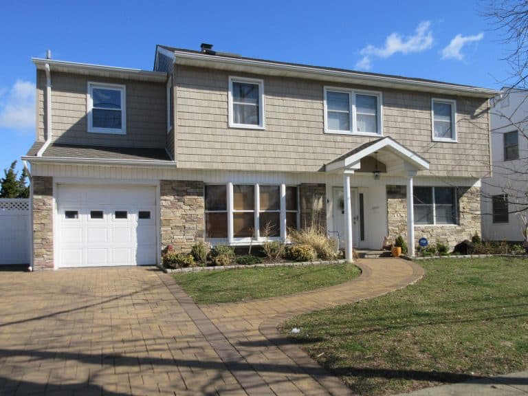 Two-story house with light brown siding and a stone facade on the lower half. The home features several windows and a white-paneled garage on the left. If you're considering this property, consider Home Inspections Long Island to ensure safety. A curved walkway leads to the front door, surrounded by a green lawn.