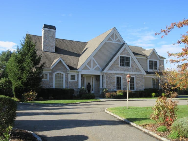 A large, two-story house with gray siding and brick accents is situated at the end of a paved driveway. It features multiple gables, a chimney, and is surrounded by well-maintained lawns and trees under a clear blue sky—perfect for those seeking thorough home inspections on Long Island.
