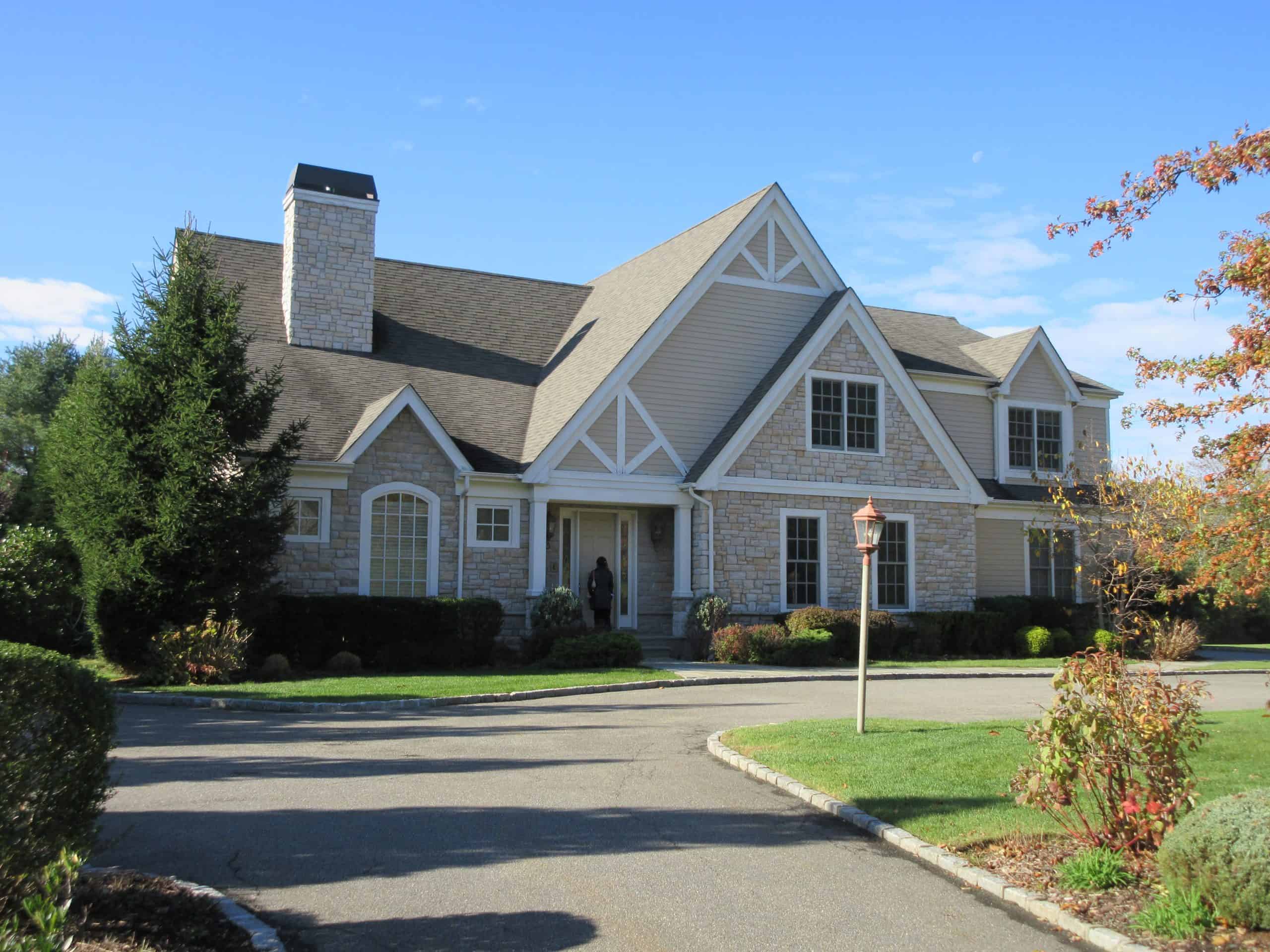 A large suburban house with beige stone and siding, peaked roofs, and multiple windows awaits a Home Inspector Long Island review. It's surrounded by a well-kept lawn and shrubs. A paved driveway leads to the entrance, where a lamppost stands near the walkway under the clear blue sky.