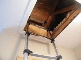 A retractable attic ladder, essential for home inspections on Long Island, is extended from a ceiling opening, leading to a dark attic space. The metal ladder features black rubber grips, contrasting the white ceiling and walls.