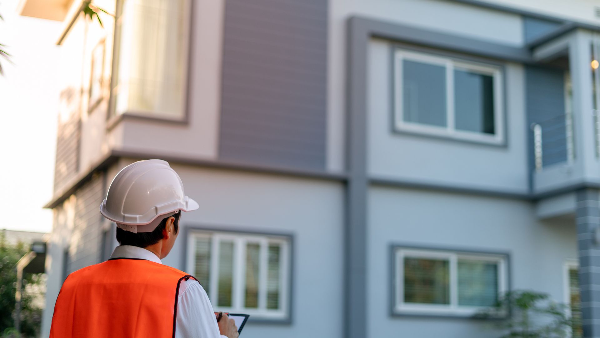 A construction worker in a white hard hat and orange safety vest holds a tablet, standing in front of a modern building with large windows. The building features gray and white walls, viewed from behind the worker, who is assisting with commercial inspections on Long Island.