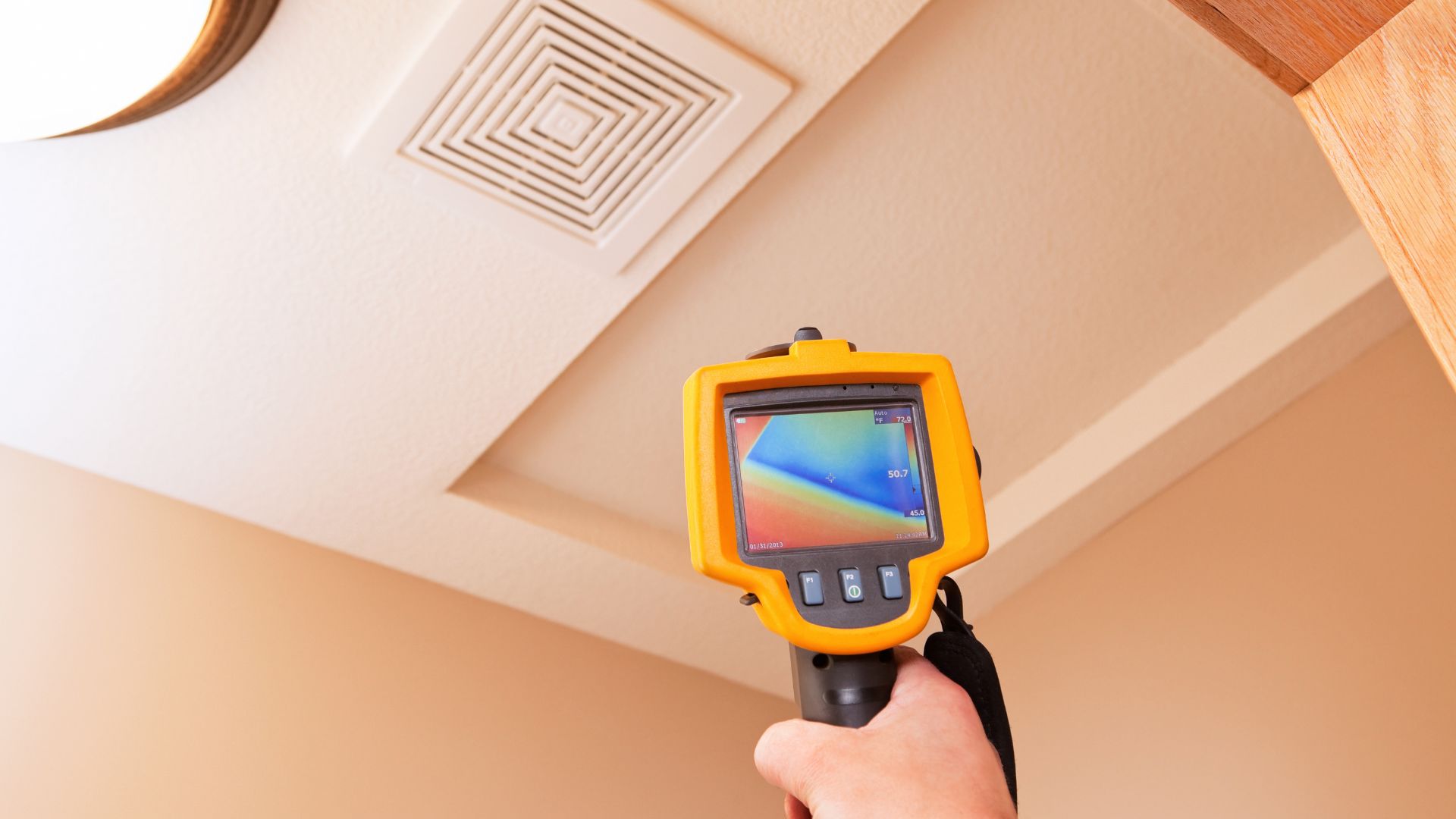 A person uses a thermal imaging camera for a home inspection in Long Island, focusing on a ceiling vent. The device displays a heat map, revealing temperature variations. The beige ceiling with its white, square air vent undergoes thorough scrutiny during this routine check.