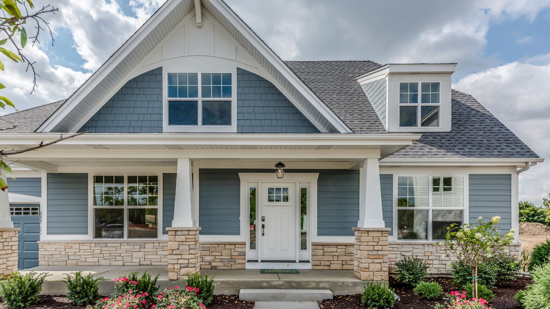 A charming blue and white house with a gabled roof, stone accents, and a covered porch. The front yard features landscaped greenery and flowers. With skies partly cloudy, it creates a welcoming atmosphere perfect for prospective buyers considering home inspections on Long Island.