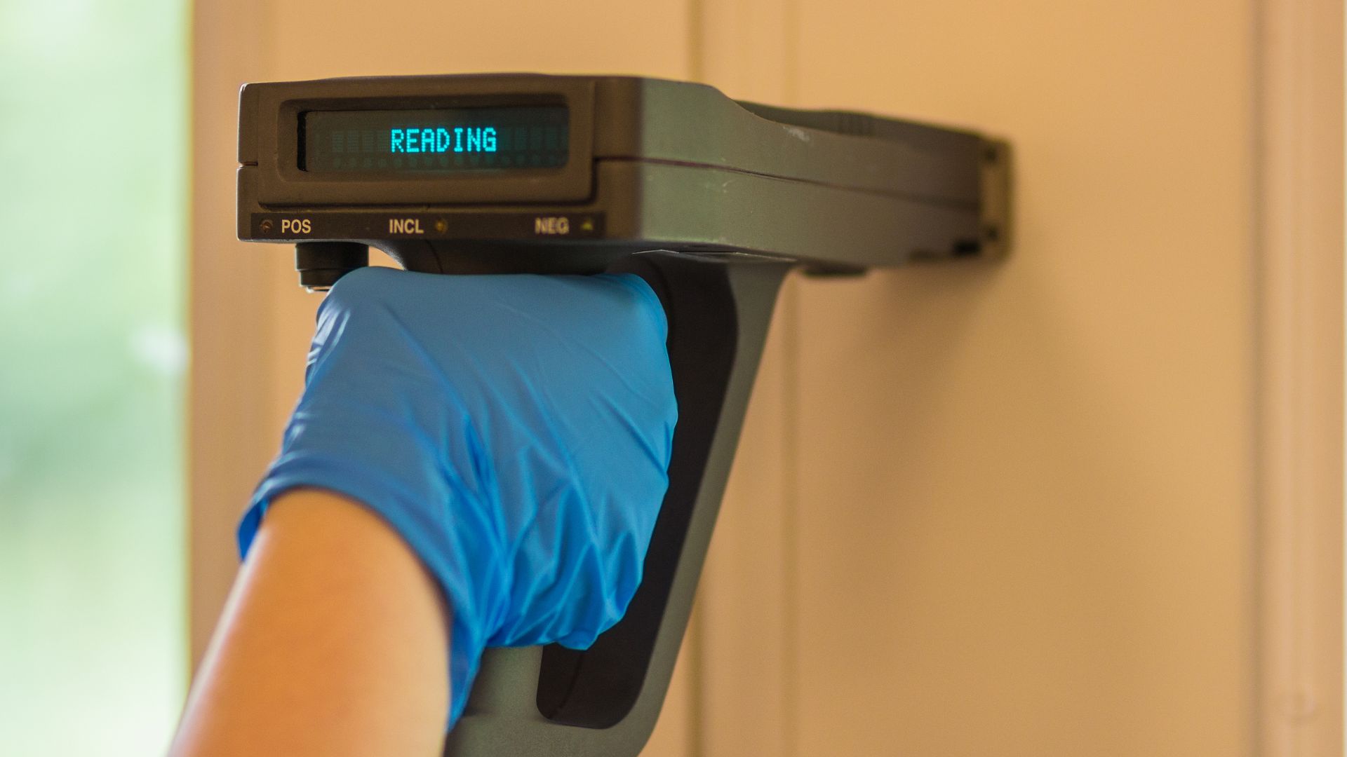 A gloved hand holds a handheld scanner against a surface, crucial for lead paint testing on Long Island. The scanner's display brightly shows the word "READING," capturing the attentive gaze of a dedicated home inspector as they ensure safety and compliance.
