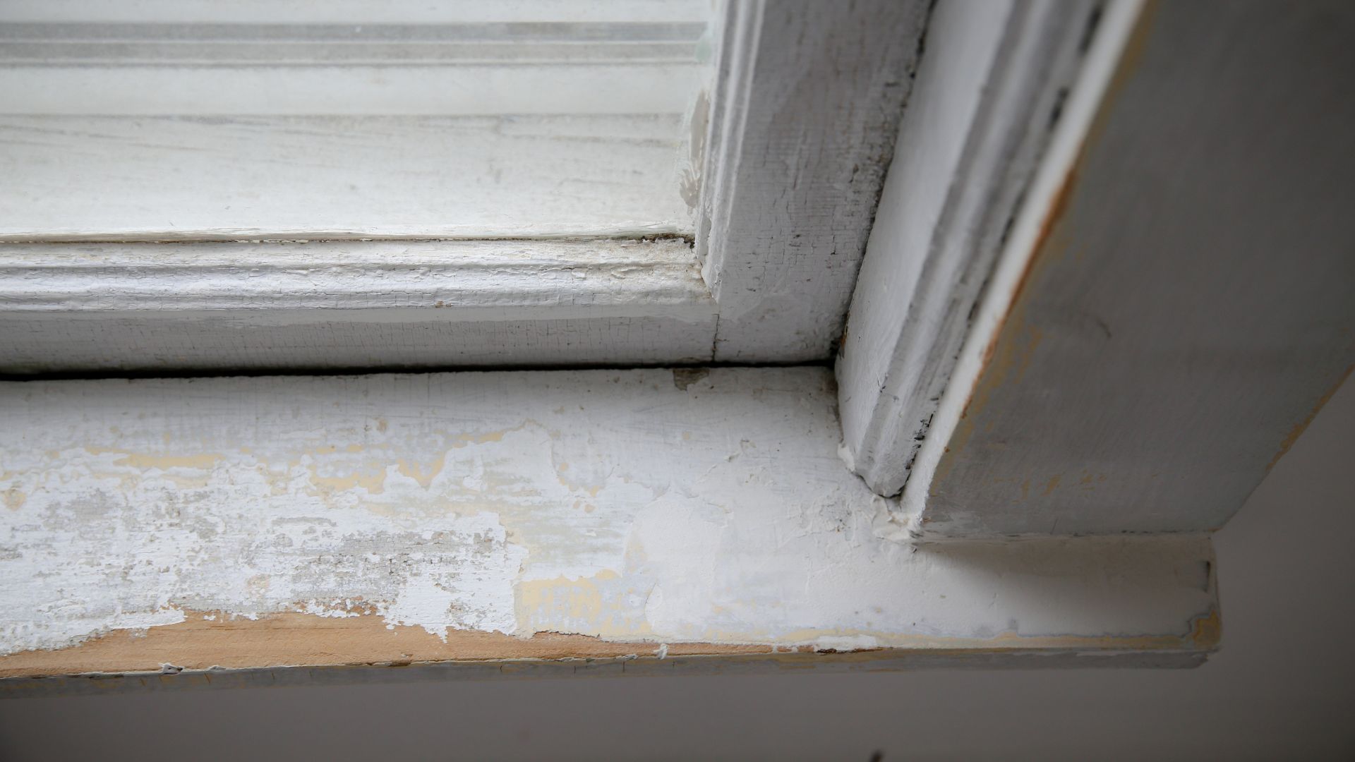 Close-up of an old wooden window frame with peeling white paint, revealing patches of bare wood. The image highlights weathering and wear, capturing a sense of age and neglect—an ideal subject for lead paint testing in Long Island homes.