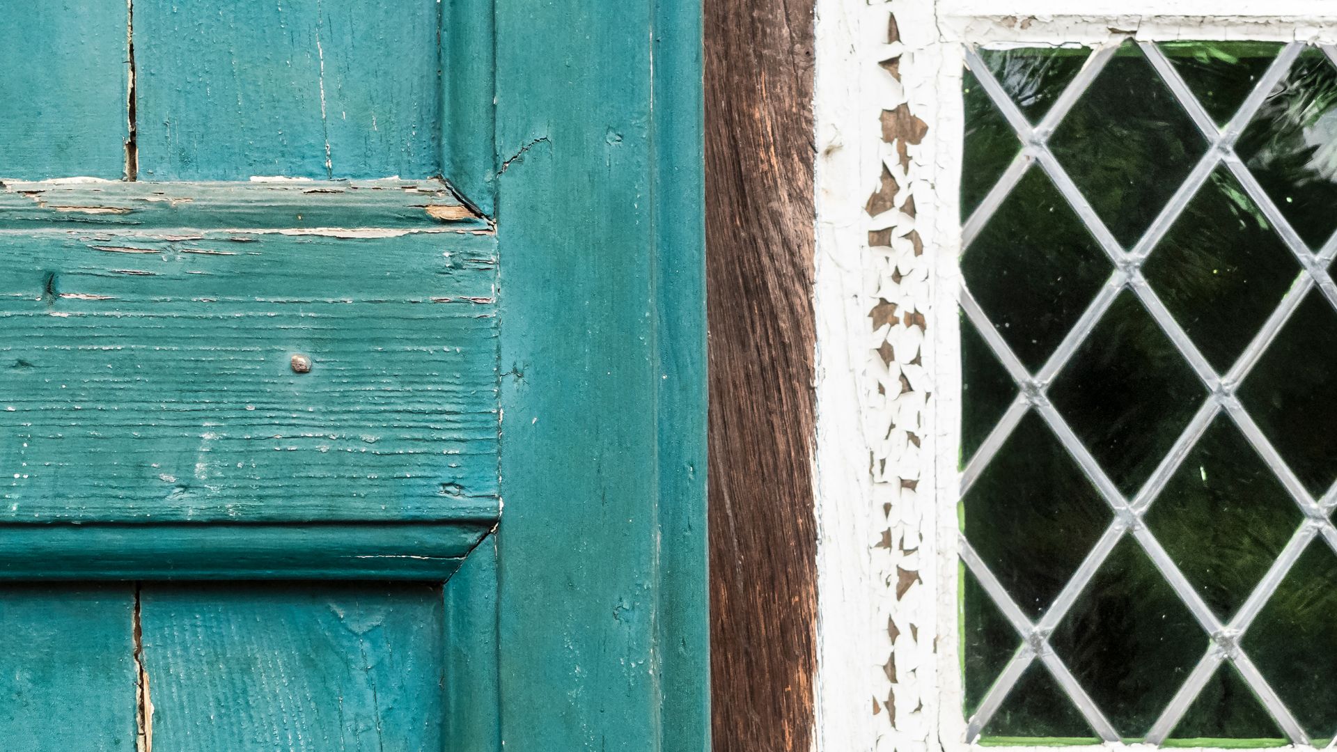 Close-up of a weathered turquoise wooden door next to a window with a white peeling frame and diamond-patterned glass. The wood grain and chipped paint evoke vintage charm, reminiscent of thorough home inspections in Long Island that assess such historic details.
