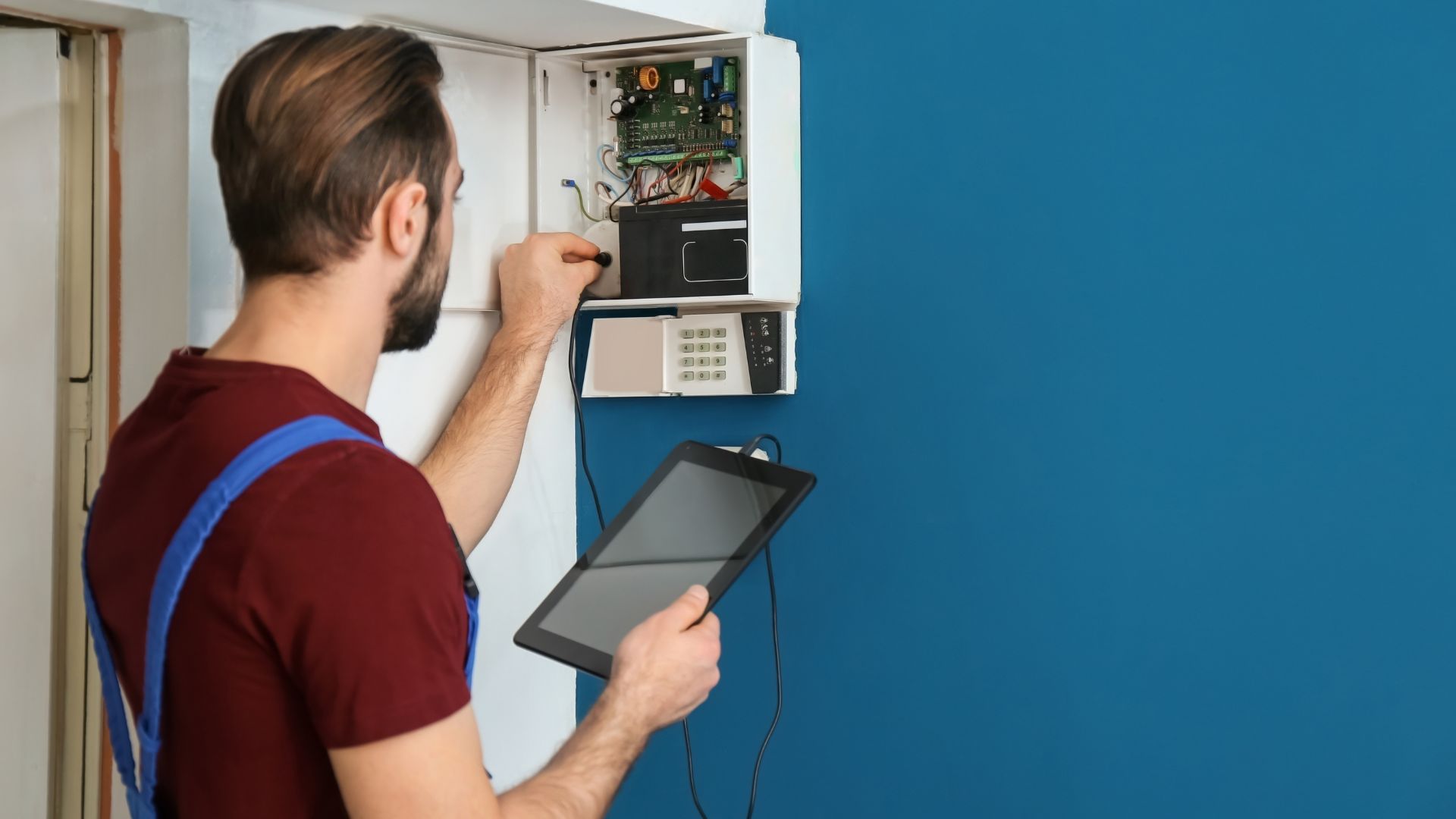 A technician with a beard and tied-back hair is adjusting a security system panel on a blue wall during a home inspection on Long Island. He holds a tablet in his other hand, wearing a red shirt and blue overalls.