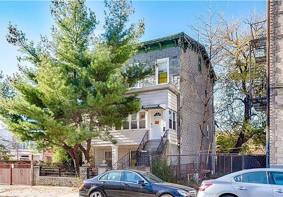 A three-story brick building with a porch, surrounded by trees, awaits its next chapter. A black car is parked on the street in front. The sky is clear and blue, and a fence encloses the property on the right side—perfect for Home Inspections Long Island's peace of mind.