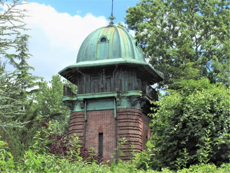 A vintage brick and copper observatory with a domed roof is nestled among lush green trees and shrubs under a partly cloudy blue sky, offering a picturesque setting that any home inspector on Long Island would appreciate.