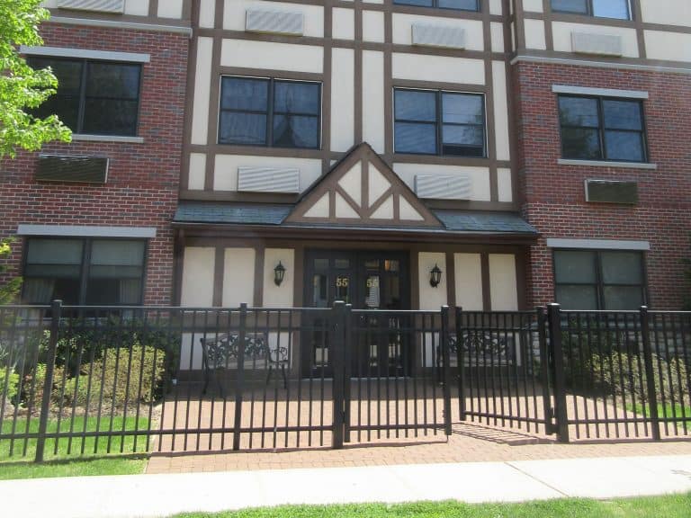 A four-story brick and stucco building with a Tudor-style facade features large windows and a black metal fence in front. Greenery lines the sidewalk, while two black lanterns flank the entrance—a prime location for commercial inspections on Long Island.