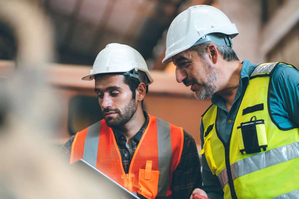 Two men in construction gear are standing together. One is wearing a white hard hat and orange vest, holding documents. The other, in a yellow vest with reflective stripes and a white hard hat, looks at the documents. They appear to be discussing a commercial inspection project on Long Island.
