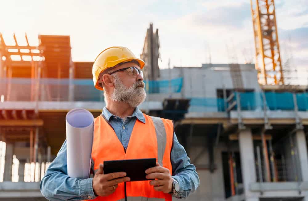 A construction worker with a beard, wearing a yellow hard hat and an orange safety vest, holds rolled blueprints and a tablet while overseeing commercial inspections on Long Island. He stands in front of a construction site at sunset.
