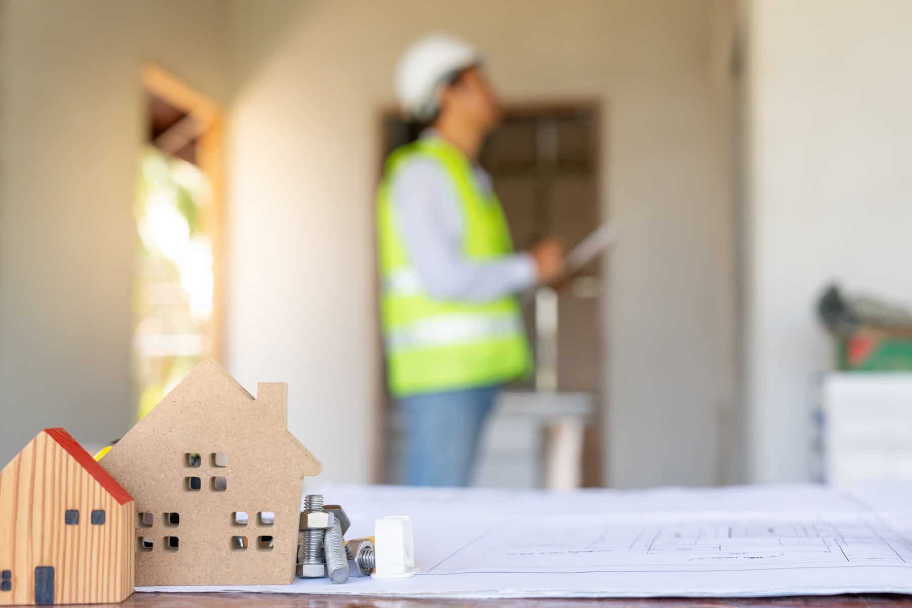 A model house and some hardware sit on a table with architectural plans. In the background, a blurred person in a reflective vest and hard hat appears to be inspecting a building site, perhaps conducting Commercial Inspections Long Island to ensure everything meets the required standards.