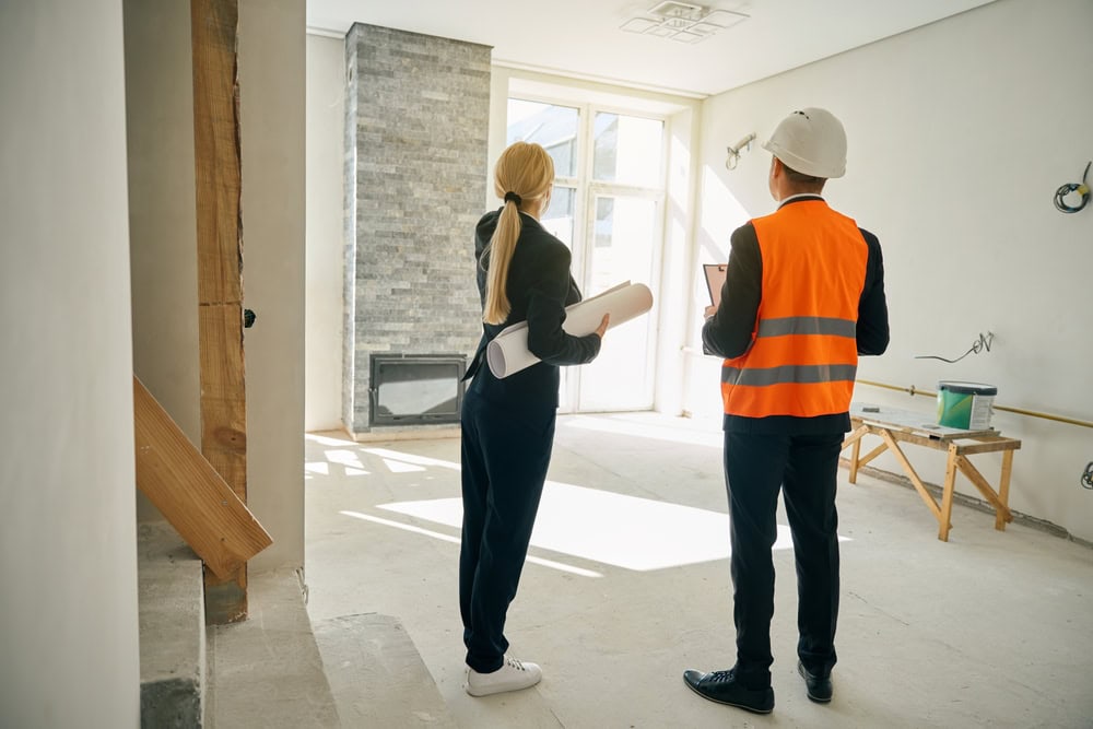 Two people stand in an unfinished room with large windows and a stone fireplace. One wears a safety vest and helmet, holding a tablet, likely for a home inspection on Long Island. The other holds architectural plans. Construction materials and tools are scattered around, hinting at ongoing work.
