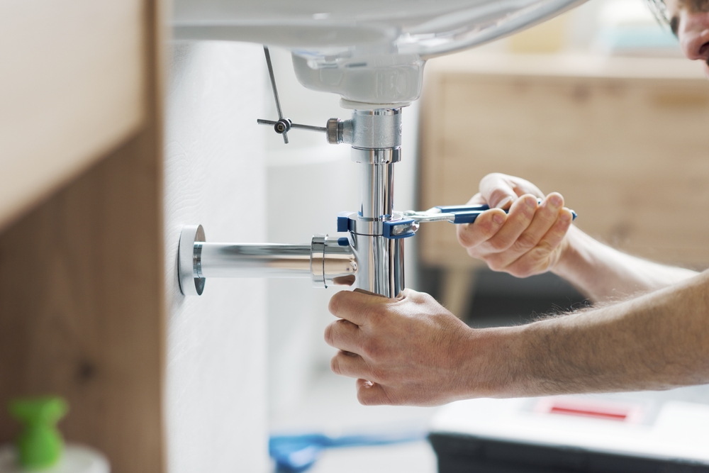 A person using a wrench to tighten a pipe connection under a bathroom sink, surrounded by various plumbing tools. This scene, akin to tasks encountered during Home Inspections on Long Island, shows hands expertly focused on the intricacies of plumbing repair or installation in progress.