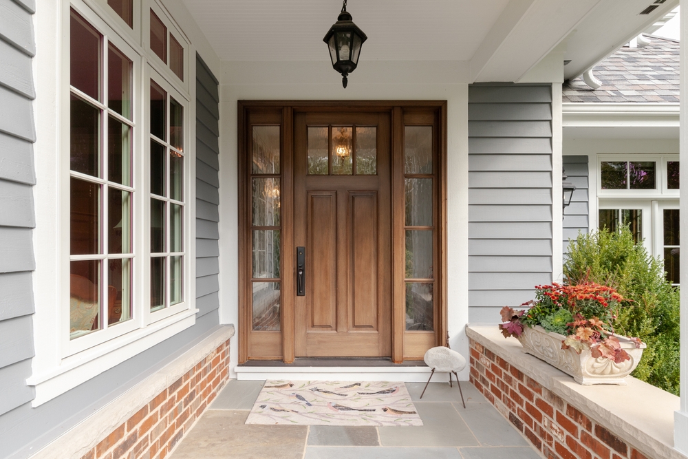 A welcoming front porch with a wooden door, sidelights, and a hanging lantern sets the stage for thorough Home Inspections Long Island. The exterior, clad in gray siding with a brick accent wall, invites you in. A patterned doormat on the stone floor and vibrant flowers in a planter complete the charming scene.