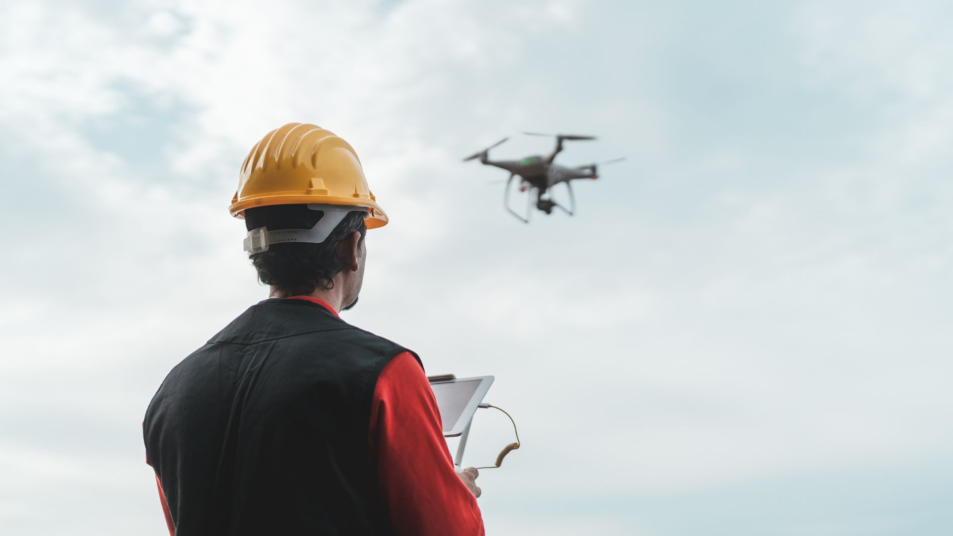 A person wearing a yellow hard hat and a black vest is operating a tablet outdoors. A drone hovers in the sky ahead, with clouds in the background.