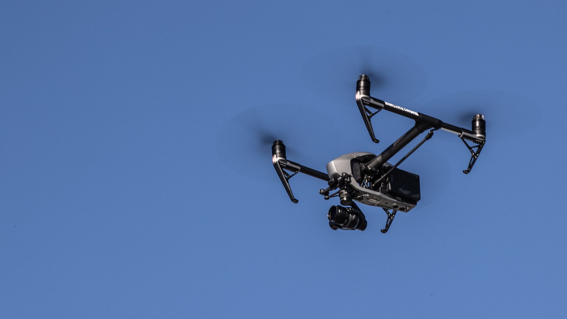 A black drone with a camera attached is flying against a clear blue sky.