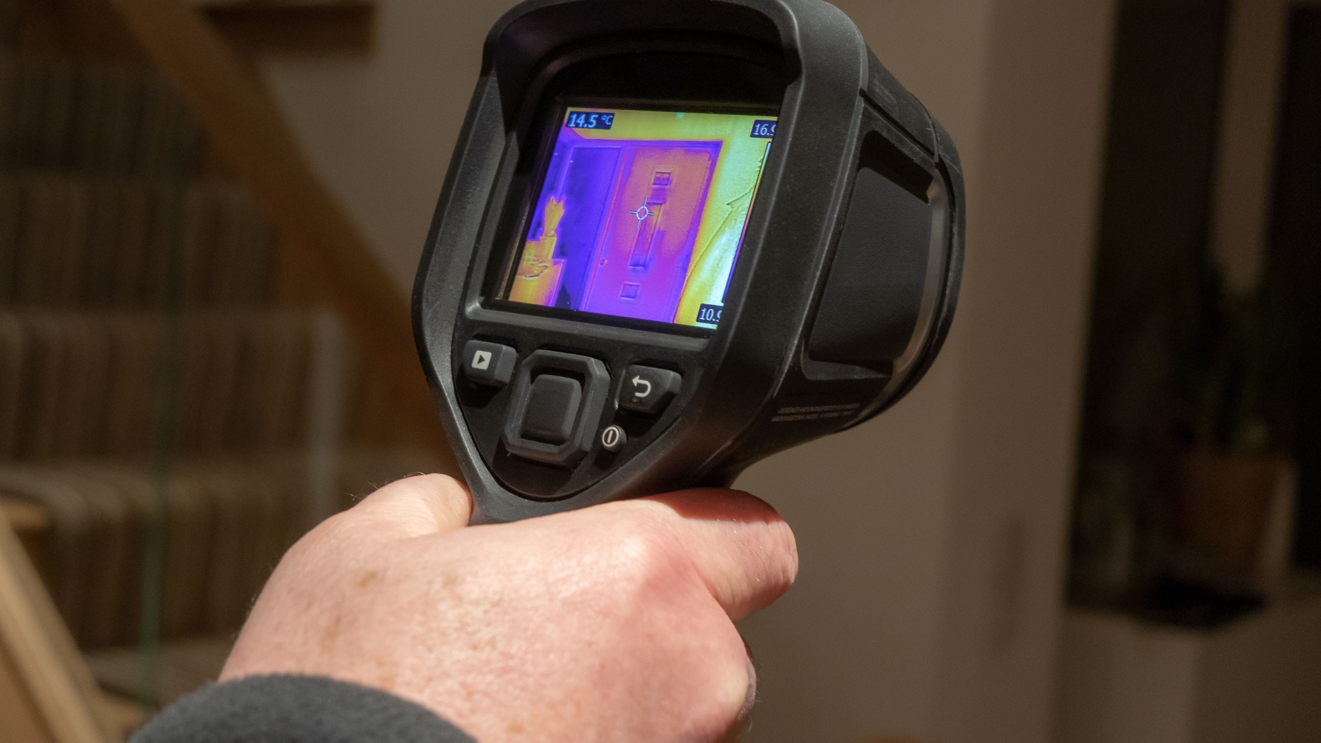 A hand holding a thermal imaging camera that displays a heat map of a segment of a staircase. The screen shows various temperature ranges in colors like purple, blue, and yellow, indicating different heat levels in the observed area.
