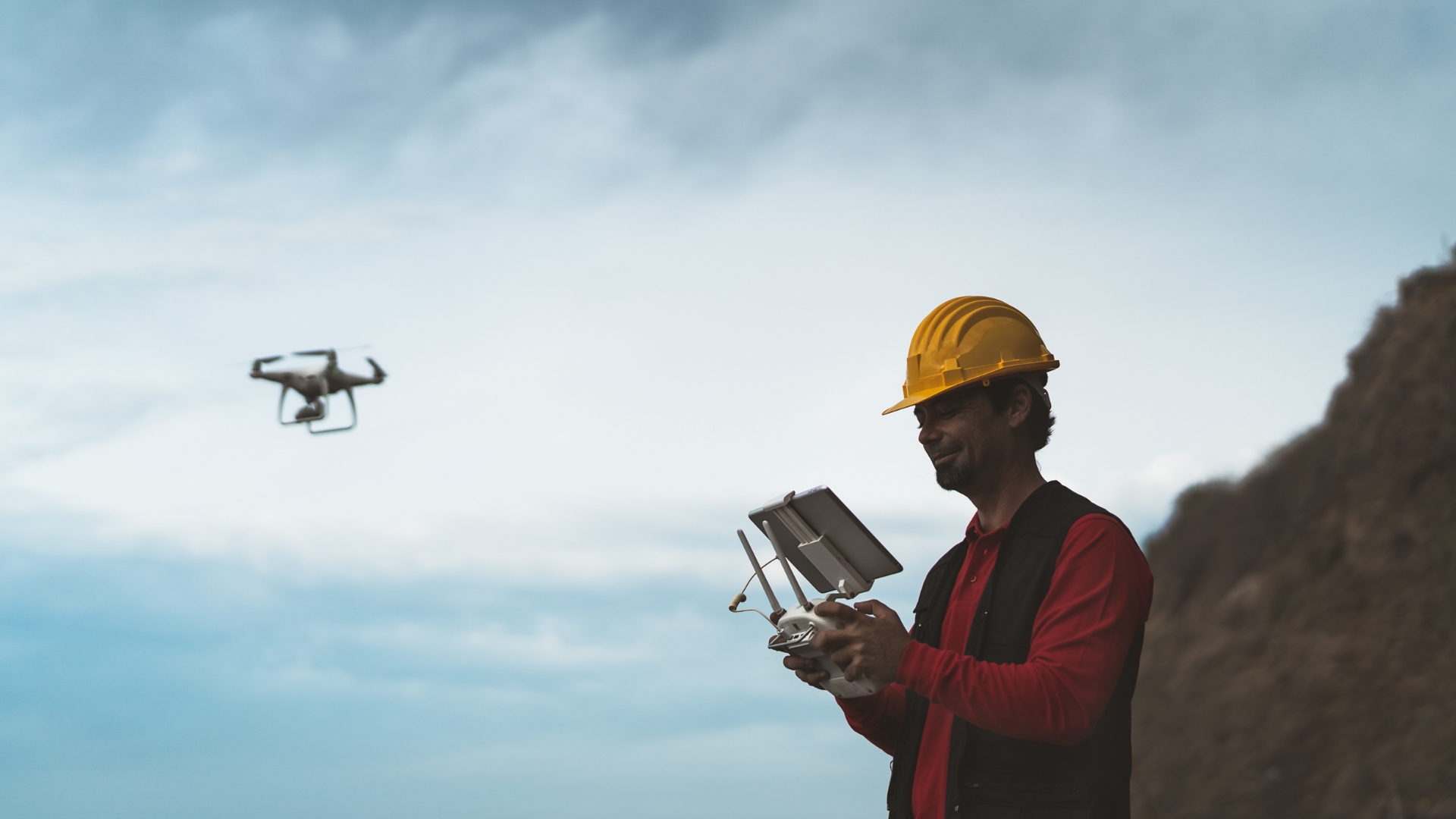 A person in a yellow hard hat operates a remote control while flying a drone in a mountainous area. The drone is visible in the sky against a backdrop of clouds and rocky terrain.