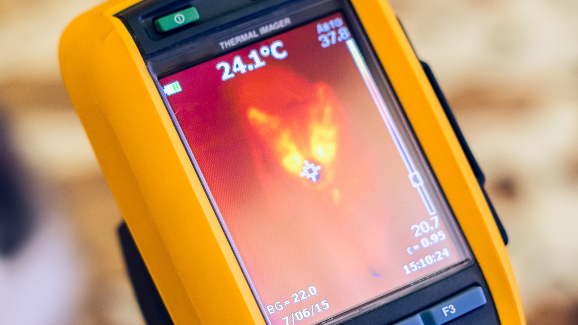Close-up of a thermal imaging device displaying a heat map in shades of orange and red. The screen shows a temperature reading of 24.1°C, with various settings and metrics visible around the edges. The device has a yellow casing.