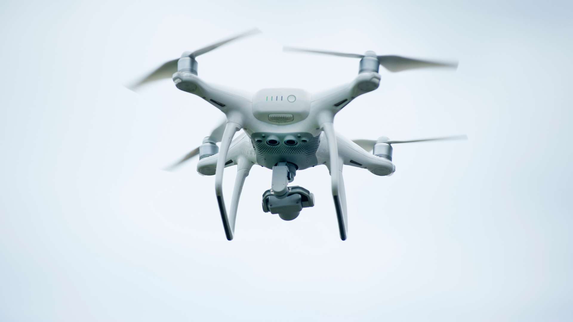 A white quadcopter drone with four propellers is flying mid-air against a light blue sky. It has a camera mounted underneath, pointed forward.
