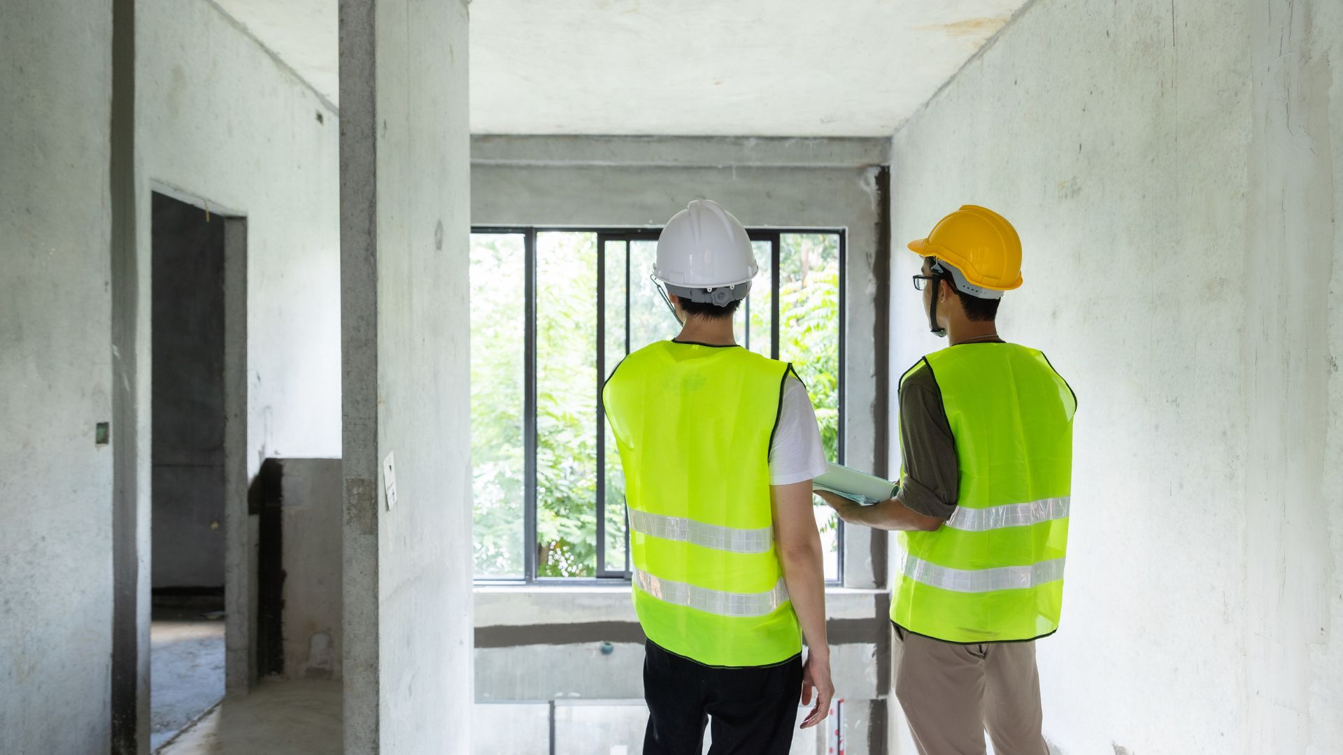 Two construction workers wearing reflective vests and helmets stand in an unfinished building, looking out at a large window. One, possibly a home inspector from Long Island, holds a rolled-up set of plans. Sunlight filters through, illuminating the empty space.