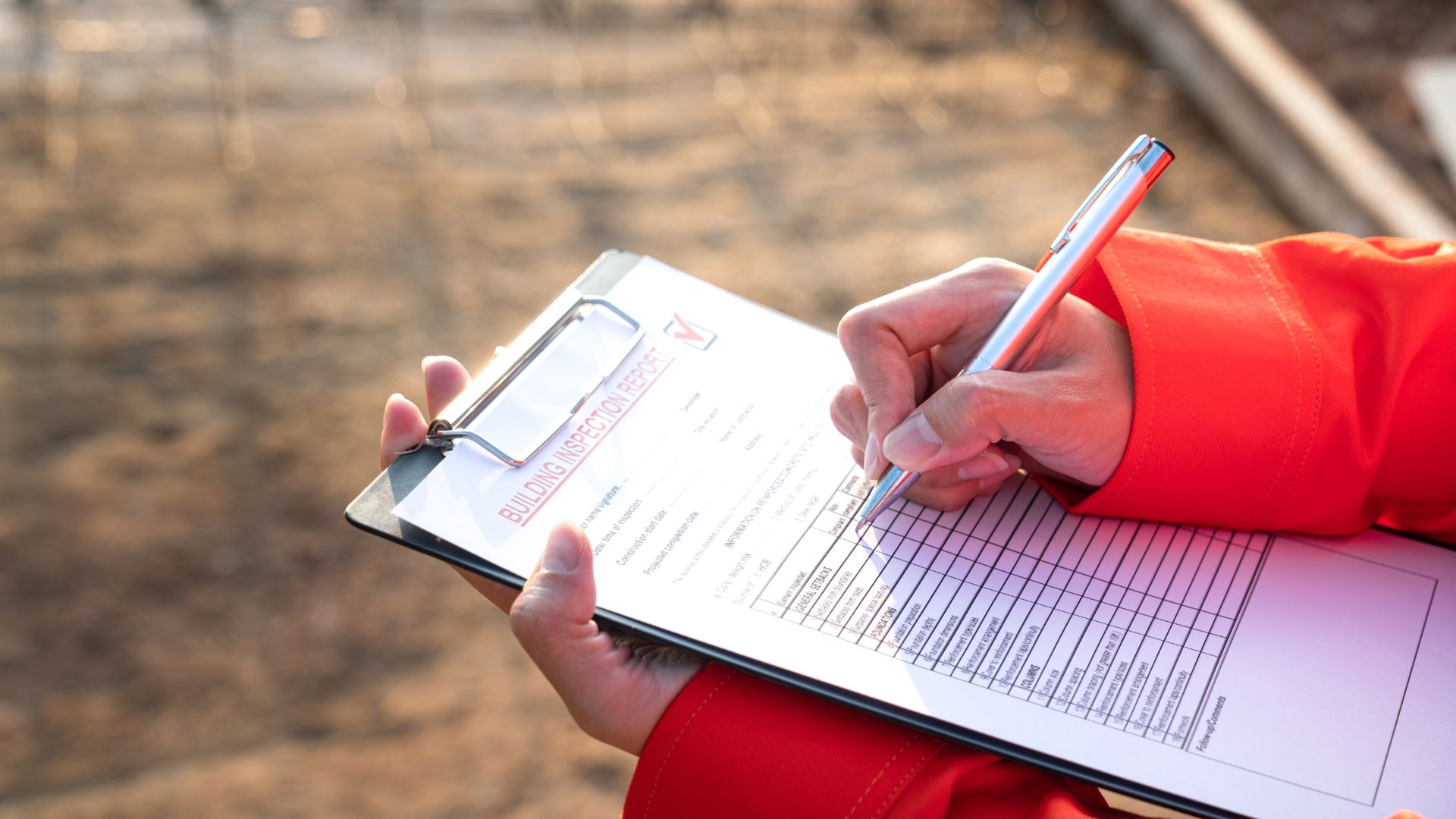A person in a red jacket holds a clipboard and pen, diligently filling out a building inspection checklist. Set against an outdoor backdrop, this scene captures the essence of home inspections Long Island, ensuring safety and compliance in residential spaces.