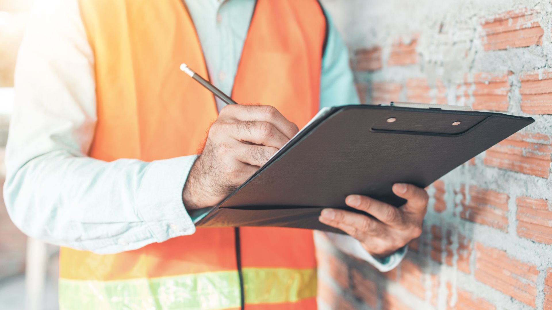 A person in a high-visibility orange vest, likely a home inspector on Long Island, holds a clipboard and writes with a pencil. Standing next to a brick wall, they seem engaged in assessing the site, emphasizing their role in meticulous home inspections across the area.