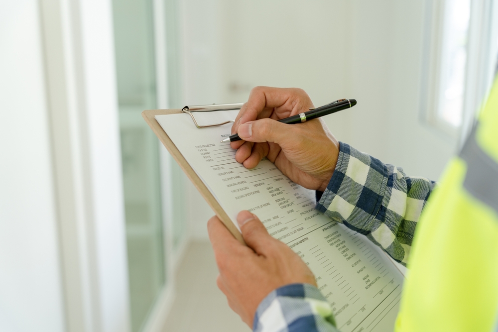 A person in a high-visibility jacket holds a clipboard with a checklist, writing with a pen. They stand in a brightly lit hallway, conducting Commercial Inspections Long Island. The focus is on the hands and clipboard, highlighting the meticulous nature of the evaluation in progress.