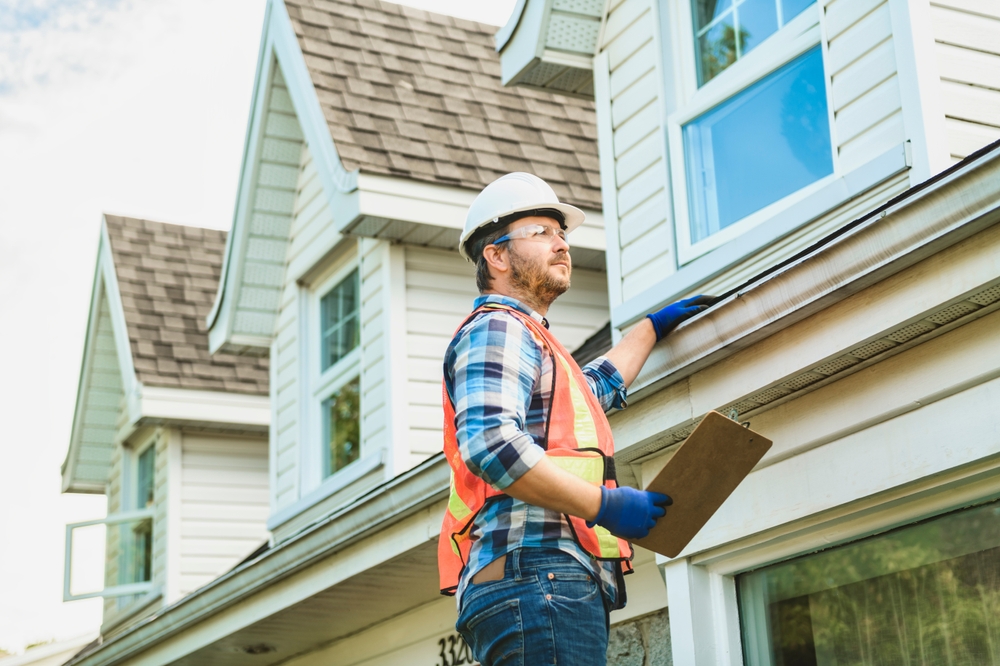 A construction worker in a hard hat and safety vest inspects the gutters of a two-story house on Long Island. As a home inspector, he holds a clipboard and wears blue gloves. The house features white siding and shingled roofing, typical for home inspections in the area.