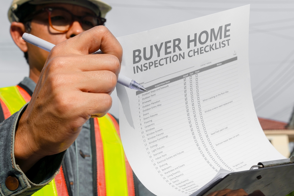 A person in a safety helmet and reflective vest holds a "Buyer Home Inspection Checklist" on a clipboard. As an expert in home inspections Long Island, they point to the checklist with a pen, emphasizing their focus on thoroughly inspecting the house. The background shows a partly visible house.